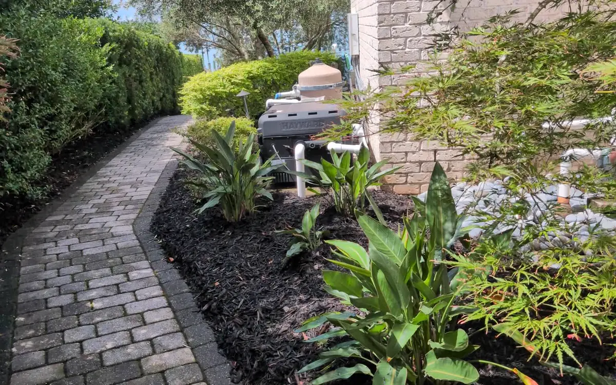 Side path past the pool gear Dark mulch bed running between a paver side path and the brick house wall
